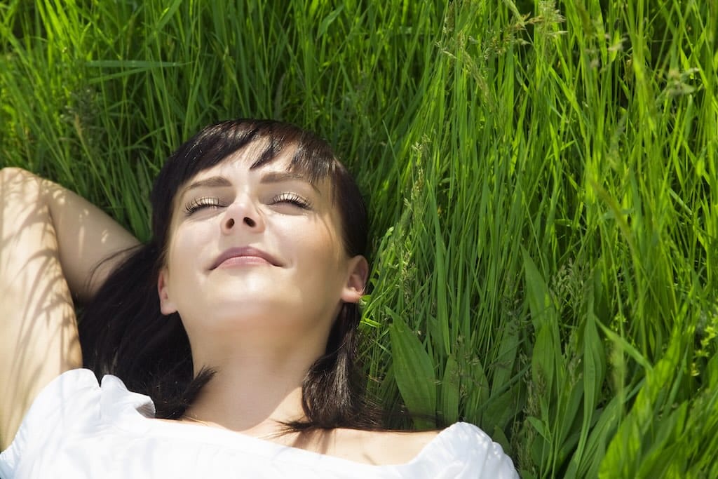 Stralende jonge vrouw ligt op het gras op een prachtige zonnige dag, ogen dicht, vredige glimlach. Diep ontspannen dankzij Hypno Atlas hypnose sessie.