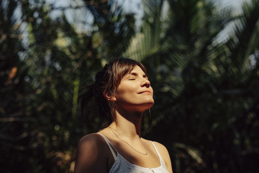 Een gelukkige jonge vrouw genietend van het warme zonlicht, mediterend met gesloten ogen, omringd door palmbomen in een tropisch openbaar park. Spiritualiteit ontwikkelen met spirituele hypnose bij Hypno Atlas.