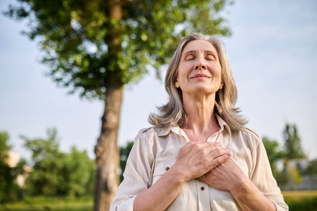 Gelukkige volwassen vrouw glimlacht met gesloten ogen, handen op de borst, ervaart innerlijke rust dankzij hypnotherapie bij Hypno Atlas.